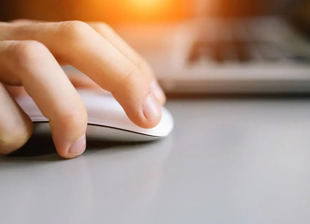 A close-up of a persons hand using a white computer mouse on a gray desk. In the background, theres a blurred laptop and a warm light shining from above, creating a soft glow.