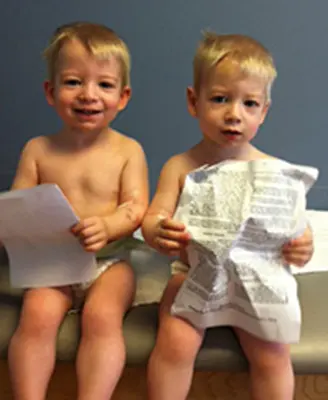 Two toddlers with blond hair sitting on a bench in diapers holding sheets of paper. One is smiling while the other looks focused. They are in a room with a blue wall background.