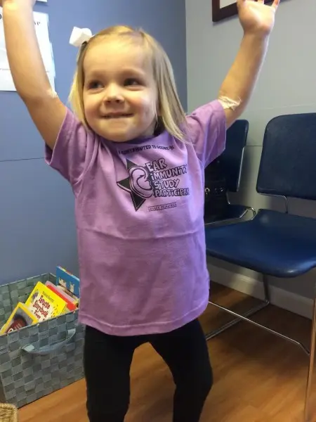 A young child with blonde hair standing with arms raised, wearing a purple T-shirt that says PEAR Immunity Study Participant. They are smiling in a room with blue chairs and a basket of books in the background.