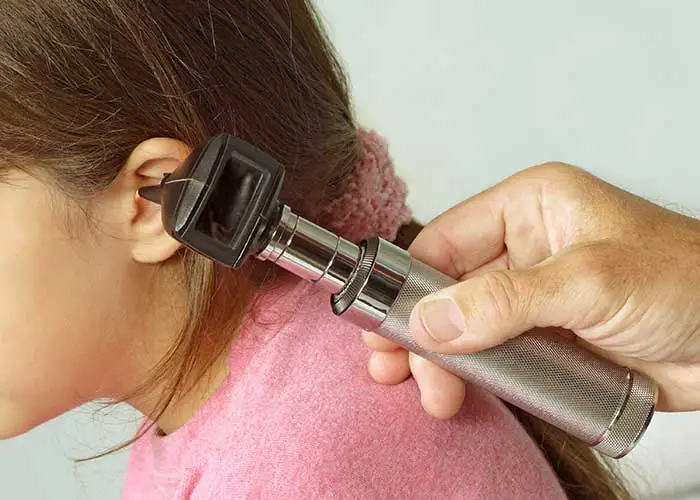 A doctor examines a young girls ear with an otoscope. The girl has brown hair and is wearing a pink shirt. The otoscope is held by an adults hand and is directed into her ear for a medical examination.