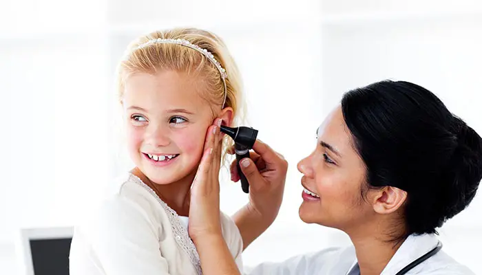 A doctor is gently examining a young girls ear with an otoscope. The girl looks happy and relaxed, with a light background in the room.