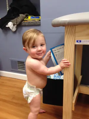 A baby in a diaper stands next to a wooden table, holding a blue and white pamphlet. The room has blue walls, a wooden floor, and a shelf with some items on it in the background.