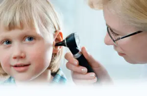 A healthcare professional examines a young childs ear using an otoscope. The child has a slight smile and short blonde hair. The adult wears glasses and focuses intently on the examination.