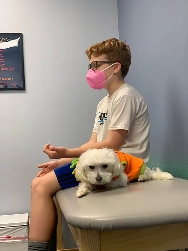 A boy wearing a pink face mask and glasses sits on an examination table in a medical room. He is wearing a white shirt and blue shorts. A small white dog in a colorful outfit rests beside him. A framed quotation hangs on the light blue wall.