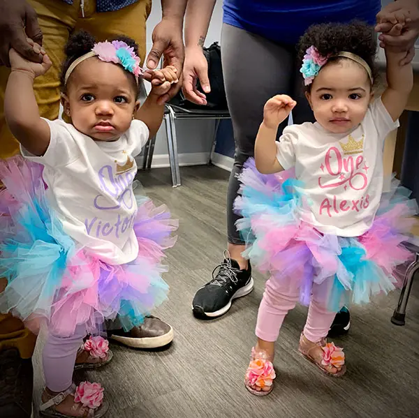 Two toddlers in colorful tutus and One shirts holding hands with adults. They have matching floral headbands and sandals. One shirt reads Victoria, the other Alexis. The room has a wooden floor and people in casual attire.