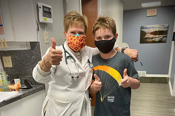 Dr. Casey gives a thumbs up while standing next to a young boy in a T-shirt and mask, who also gives a thumbs up. .