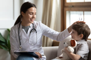 A smiling female doctor with a stethoscope gently pats a young boy’s head. The boy sits on a couch, holding a stuffed dog and smiling. They appear to be in a comfortable, well-lit office or home setting.