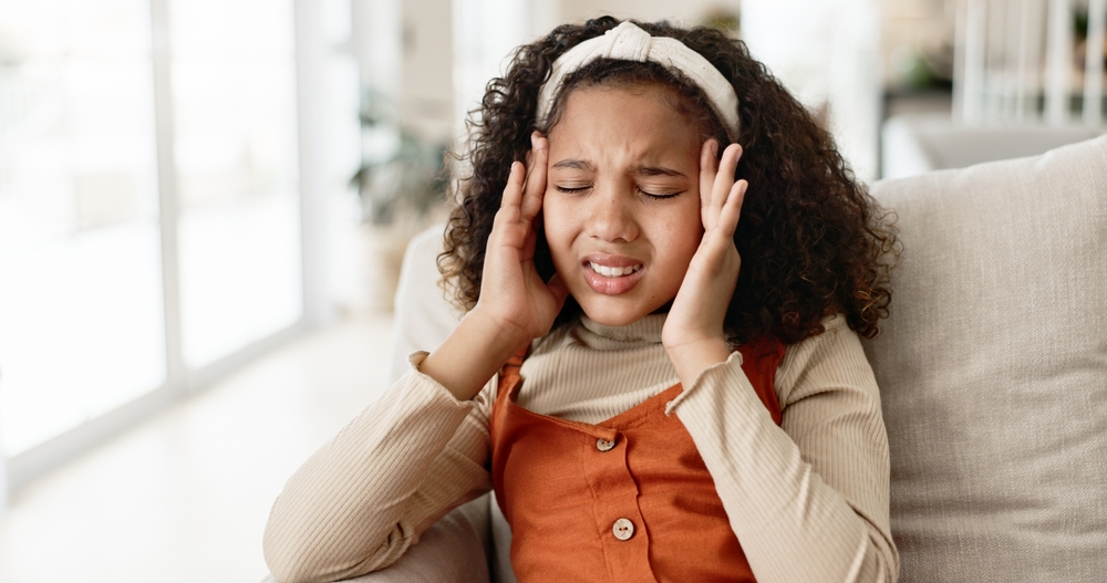 A young girl with curly hair and a headband sits on a couch, eyes closed and hands on her temples, appearing to be in pain or discomfort. She is wearing a beige turtleneck and an orange dress.
