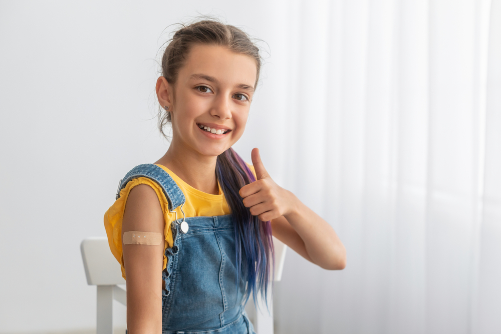 A smiling girl wearing denim overalls shows a thumbs up while sitting on a chair, with a bandage on her upper arm, suggesting she just received a vaccination. White curtains are in the background.