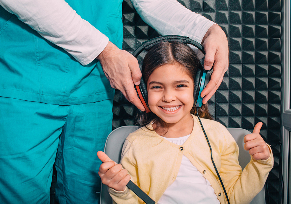 A smiling girl sits in a soundproof room wearing large headphones, giving two thumbs up. An adult in medical scrubs adjusts the headphones on her head, suggesting a hearing test or audiology exam.