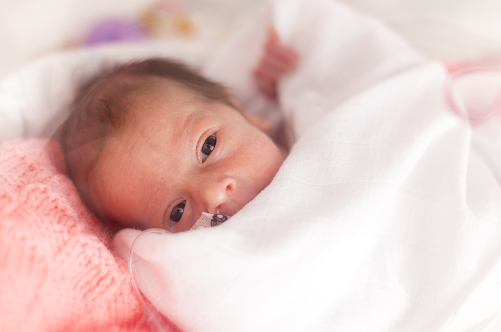 A newborn baby wrapped in a white blanket lies on a pink knitted blanket, looking up with wide eyes. The baby appears cozy and calm in a soft, gentle setting.