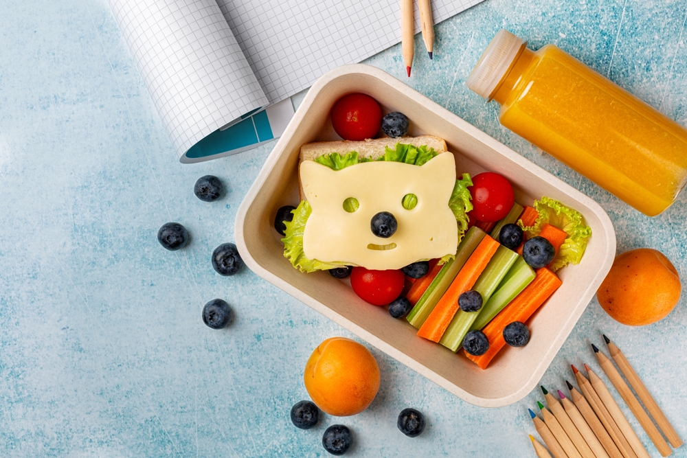 A lunchbox with a sandwich decorated as a cat face using cheese, peas, and nori, surrounded by lettuce, tomatoes, celery, carrots, and blueberries. Nearby are colored pencils, an open notebook, a juice bottle, and two mandarins.