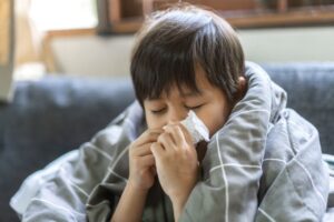 A young child wrapped in a gray blanket sits on a couch with eyes closed, blowing their nose into a tissue, appearing to be sick or have a cold—a relatable scene in any flu guide for parents.