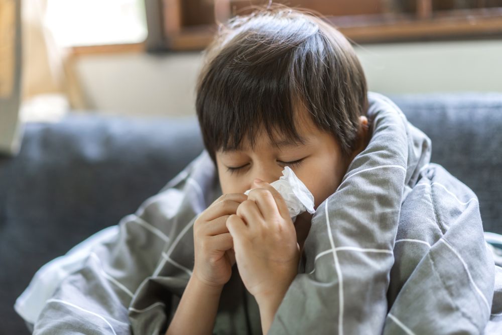 A young child wrapped in a gray blanket sits on a couch with eyes closed, blowing their nose into a tissue, appearing to be sick or have a cold—a relatable scene in any flu guide for parents.