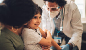 A young child sits on an adult’s lap, smiling, while a healthcare professional in protective gear prepares to give the child a vaccine or medical shot.