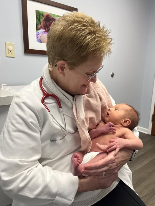 A smiling healthcare professional in a white coat and red stethoscope holds a newborn baby wrapped in a pink blanket while sitting in a medical office. A photo hangs on the wall in the background.